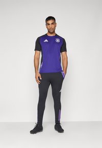 Maglietta da calcio viola con maniche nere e logo. Leggings neri aderenti con strisce viola alla caviglia. Indossati con scarpe da calcio nere.