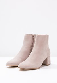 Bottines en suède beige clair avec des talons carrés et des zips sur le côté, présentées sur une surface blanche et un fond blanc.