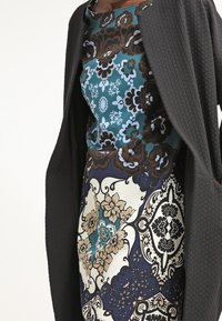 Mörkgrå texturerad kappa över en blommönstrad klänning med blå, brun och krämfärgade nyanser med intrikata mönster och accenter.