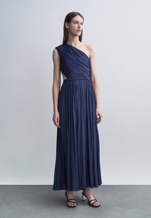 Femme portant une robe longue plissée bleu marine à une épaule et des talons noirs à brides, debout devant un fond gris clair uni.