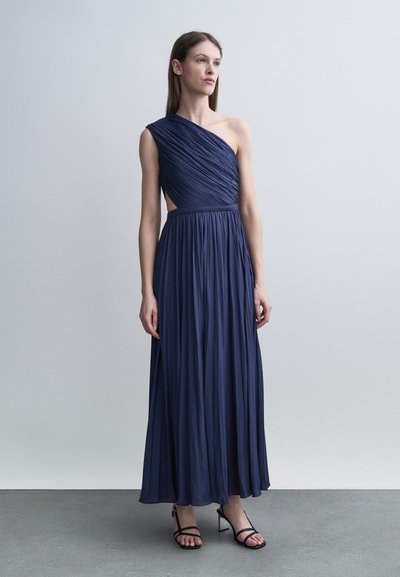 Femme portant une robe longue plissée bleu marine à une épaule et des talons noirs à brides, debout devant un fond gris clair uni.