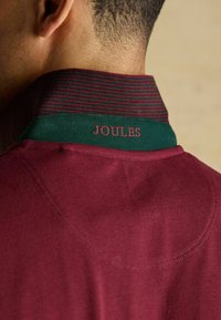 Merepunane polo särk, millel on roheline ja burdgundi triibuline krae, koos roosa "JOULES" tikandiga krael. Pehme tekstuur.