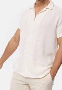 Short-sleeve, light beige shirt with a textured weave, button closure, and a relaxed fit. Features a notched collar and straight hem.