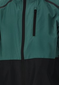 Veste verte et noire avec fermeture à glissière, tissu texturé, et une poche zippée sur la poitrine, avec des détails de surpiqûres en contraste.