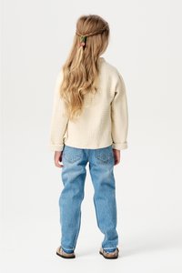 Jasno beżowy, teksturowany bawełniany top z podwiniętymi rękawami, w połączeniu z luźnymi niebieskimi dżinsami i beżowymi sandałami, widok od tyłu.