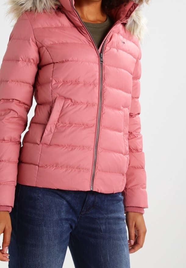 Femme portant une veste matelassée rose poudré zippée avec une capuche bordée de fourrure et des poches latérales, associée à un jean bleu foncé.