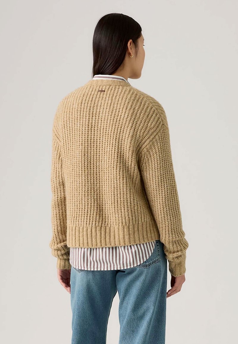 Maglione beige lavorato a maglia con un motivo testurizzato, orlo a coste e maniche lunghe, indossato sopra una camicia a righe bianca e marrone.