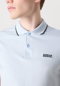 Cămașă polo de un albastru deschis, cu un placket cu nasturi, o textură moale și un guler cu accente bleumarin. Afișează logo-ul "Barbour International".