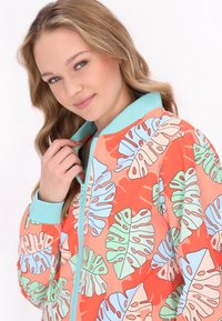 Giacca leggera con zip, caratterizzata da uno sfondo corallo vivace e grandi motivi di foglie verdi e blu. Accentuata con un collo di colore menta.