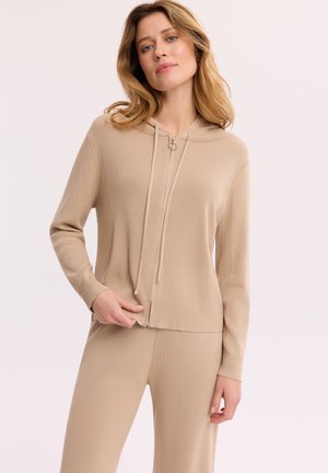 Kobieta o blond włosach do ramion, ubrana w beżową bluzę na zamek i dopasowane spodnie na jednolitym jasnym tle.