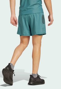 Persoon in teal sportshorts en top, grijze sokken en zwarte hardloopschoenen, staand op een lichte achtergrond.