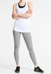 Camiseta sin mangas blanca sobre un sujetador deportivo negro, combinada con leggins grises con acentos negros. La modelo lleva zapatillas deportivas negras.