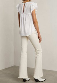 Vit, texturerad blus med volanger vid ärmarna och nyckelhål i ryggen; kombinerad med vita utsvängda jeans och svarta klackstövlar med dekorativa detaljer.