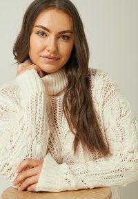 Maglione bianco lavorato a maglia con collo alto, intricate trame a treccia e pizzo, e polsini a costine. La mano del modello è appoggiata su una superficie in legno chiaro.