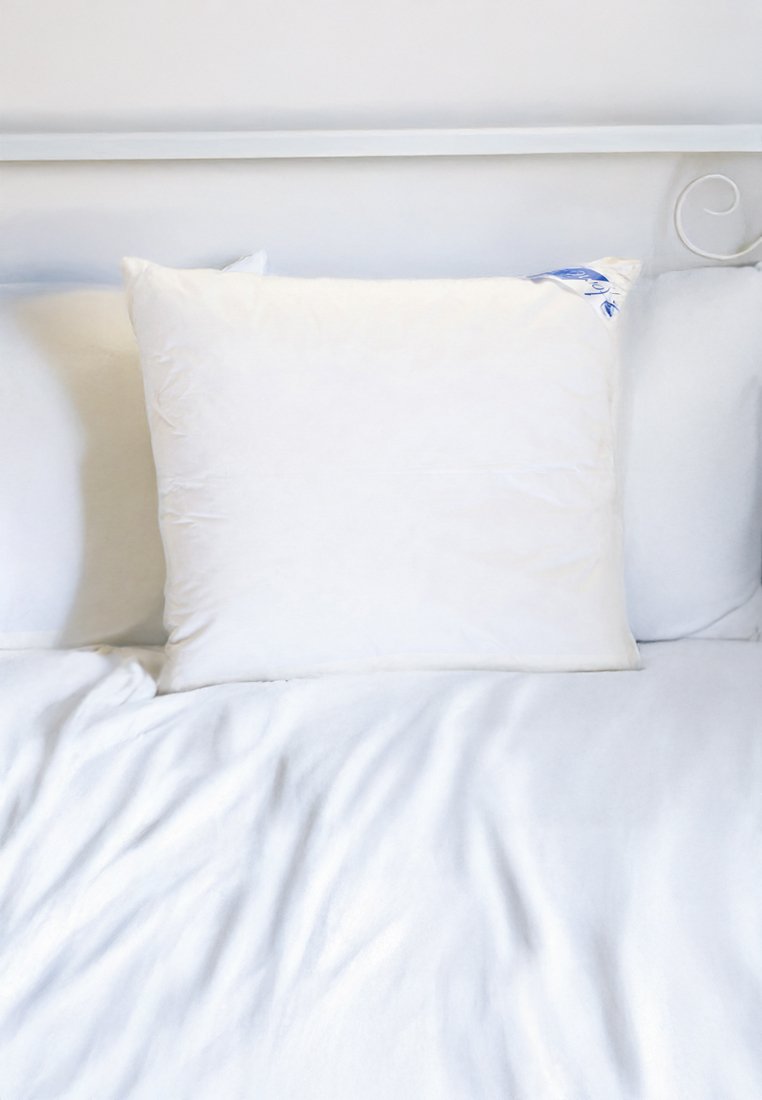 Ein weißes Kissen auf einem weichen weißen Bettüberwurf. Das Kissen hat eine glatte Textur und ein blaues Etikett in einer Ecke, vor einem neutralen Hintergrund.