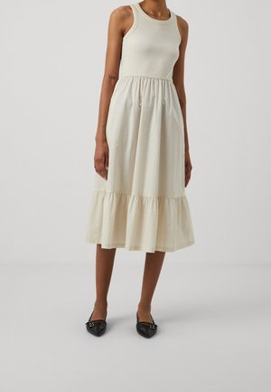 Mannequin féminine portant une robe midi crème sans manches avec une jupe plissée et des ballerines noires à bouts pointus, posant devant un fond uni.