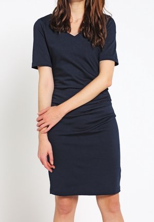Woman wearing a fitted navy blue short-sleeve dress with a V-neck, standing with arms crossed lightly in front of her.