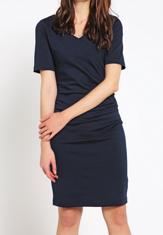 Femme portant une robe ajustée bleu marine à manches courtes avec un col en V, debout, les bras légèrement croisés devant elle.