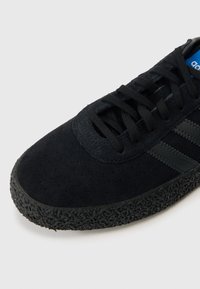 Ανδρικό αθλητικό παπούτσι σε μαύρο χρώμα, με σουέντ επάνω μέρος με διάτρητη υφή, μαύρη καουτσούκ σόλα και τρεις σκουρόχρωμες γκρι ρίγες. Ορατή μπλε ετικέτα με λογότυπο.