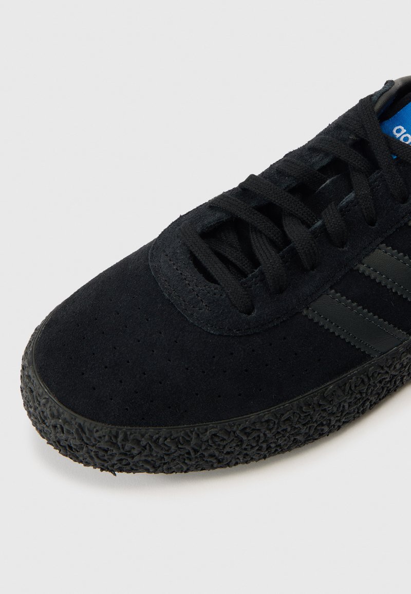 Schwarzer Sportschuh für Herren mit einem Obermaterial aus Wildleder mit perforierter Textur, schwarzer Gummiaußensohle und drei dunkelgrauen Streifen. Ein blaues Logoetikett ist sichtbar.