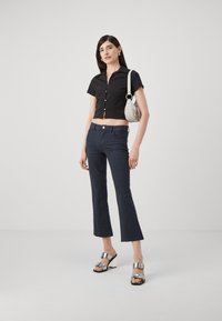 Zwarte cropped button-up shirt, gestructureerde donkerblauwe flared broek met een patroon, en zilveren hakken sandalen. Een kleine witte handtas accentueert de look.