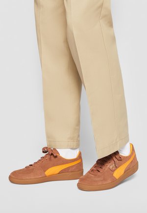 Sneaker in suede marrone con accenti e lacci gialli. Altezza media, con una finitura testurizzata e suola in gomma. Abbinati a pantaloni beige.