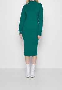 Robe ajustée à manches longues de couleur bleu sarcelle avec un col montant et une texture côtelée, associée à des bottines blanches chevilles. Design simple et épuré.