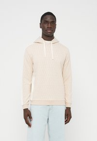 Svetlo bež pulover s kapuco z diamantnim vzorcem, belimi vrvicami za zategovanje, rebrastimi manšetami in robom. Kombiniran s svetlo modrimi kavbojkami. Mehka tekstura, priložnostna prileganje.