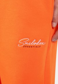 Orange Baumwoll-Jogginghosen mit weißen gestickten Text "Smilodox ESSENTIALS" an der Seite. Sanfte Textur mit enger Passform.