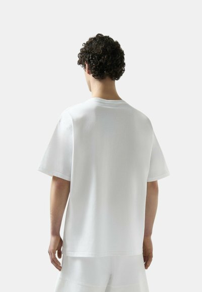 Uomo con capelli ricci scuri che indossa una maglietta bianca a maniche corte e pantaloncini bianchi, visto di spalle su uno sfondo chiaro.