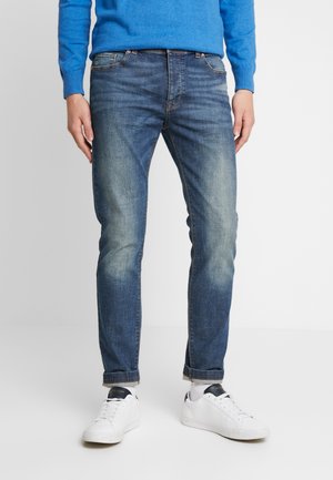 Mężczyzna ubrany w wyblakłe, niebieskie, obcisłe dżinsy, białe trampki i niebieski sweter stoi na tle jednolitego jasnego tła.