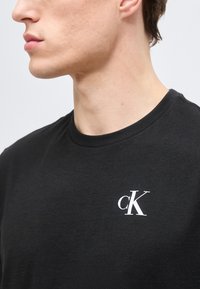 Κοντινό πλάνο ενός ατόμου που φοράει μαύρο μπλουζάκι με λευκό λογότυπο "cK" στο αριστερό στήθος, πάνω σε ανοιχτόχρωμο φόντο.