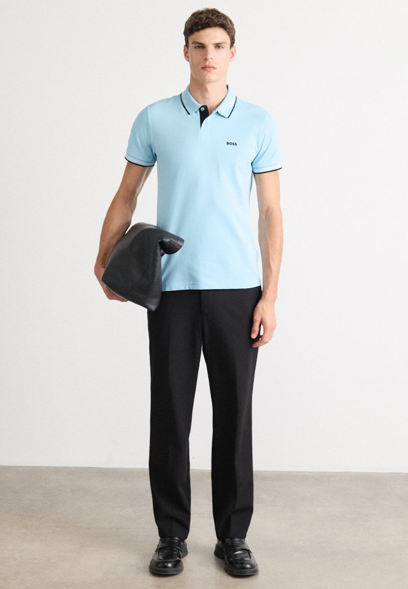 Polo shirt bleu clair avec des accents et un col noir, pantalon noir, chaussures noires brillantes, et un sac en cuir noir tenu dans la main gauche.