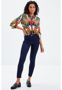 Blusa multicolore e fantasia con maniche arrotolate, abbinata a jeans skinny blu scuro e scarpe nere con finitura texturizzata.
