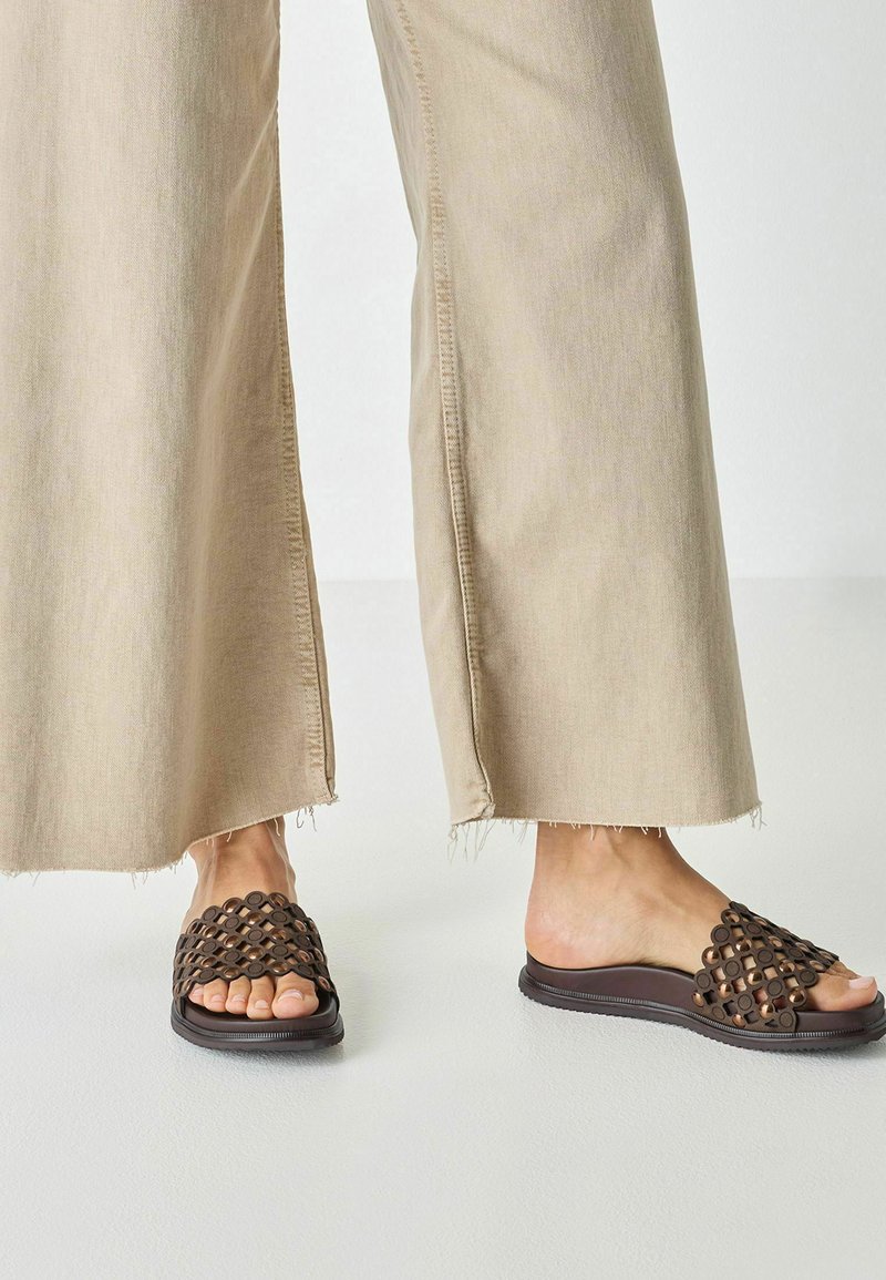 Sandales slides tissées marron avec découpes circulaires, semelle plate et texture lisse. Portées avec un pantalon beige à jambes larges avec ourlet effiloché.
