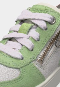 Κοντινή λήψη πράσινου και λευκού αθλητικού παπουτσιού με λευκά κορδόνια και μεταλλικό φερμουάρ στο πλάι, που δείχνει υφές υλικών και λεπτομέρειες ραφής.