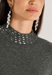 Frau mit langen Ohrringen aus Kristalltropfen und einem dunkelgrauen Strickpullover mit Kristallverzierungen am Kragen.