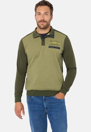 Mann mit kurzen dunklen Haaren und Bart, der einen olivgrünen Pullover mit langen Ärmeln und blaue Jeans trägt, steht mit einer Hand in der Tasche.