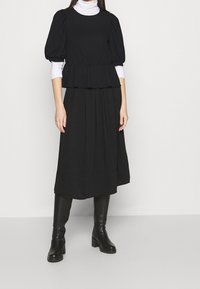 Černý peplum top s nafouknutými rukávy, vrstvený přes dlouhou černou sukni. Doplněný černými koženými botami po kolena. Jednoduchá, texturovaná tkanina.