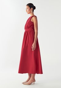 Sieviete sarkanas bezpiedurkņu midi kleitā un dzeltenīgās kaķenēs stāv profila pozā uz viendabīga baltā fona.