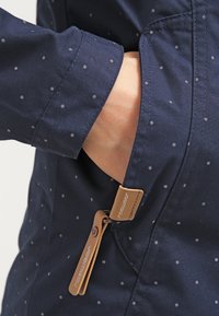 Mörkblå jacka med vita prickar, med en sidoficka och en beige dragkedjeanordning med varumärkesdetalj. Slät tygtextur.