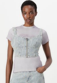 Denim crop top featuring a light blue wash, front zipper, and wide straps. Worn over a fitted, light purple ribbed shirt.