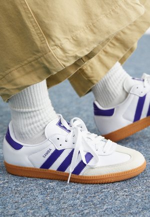 Białe i fioletowe buty Adidas Samba z gumowymi podeszwami noszone z białymi prążkowanymi skarpetkami i beżowymi spodniami na wykładzinie.