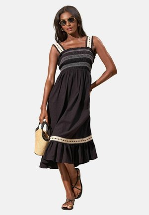 Nő fekete midi ruhát visel fehér csipkedíszítéssel és fodros szegéllyel, fekete szandált, napszemüveget, és szalmatáskát hord.