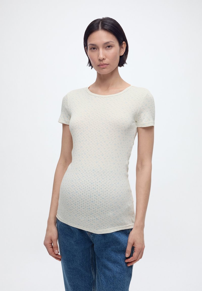 Νεαρή γυναίκα με κοντά σκουρόχρωμα μαλλιά, φοράει εφαρμοστή ανοιχτό μπεζ μπλούζα με σχέδια και κοντά μανίκια, καθώς και μπλε τζιν, μπροστά σε απλό λευκό φόντο.