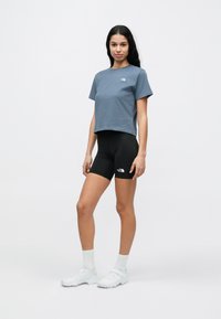 Donna in piedi che indossa una T-shirt corta blu, pantaloncini neri da biker, calzini bianchi e scarpe da ginnastica bianche su uno sfondo uniforme.