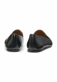 Een paar zwarte leren platte loafers, gezien van achteren op een witte achtergrond, met een gladde afwerking en minimale stikdetails.