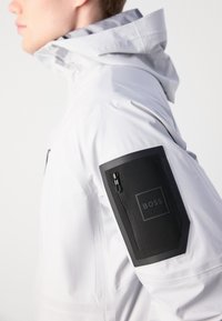 Bărbat îmbrăcat cu o jachetă albă cu glugă, având un patch negru cu fermoar pe mâneca superioară, etichetat "BOSS."