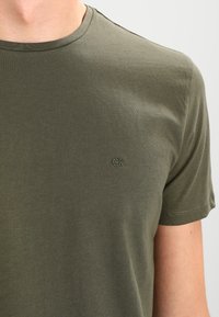 Mężczyzna ubrany w oliwkową koszulkę z krótkim rękawem i okrągłym dekoltem, z małym wyszywanym logo "CK" po lewej stronie klatki piersiowej.