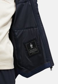 Giacca in pile beige con fodera blu scuro, dotata di una tasca interna con un'etichetta nera, apertura con zip e texture liscia.